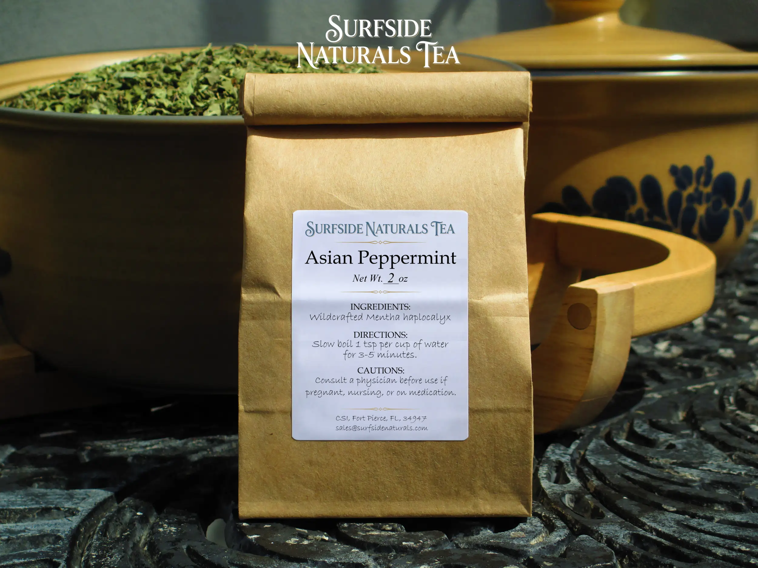 Tin-tie kraft package sitting on a black wrought-iron table with a white label that shows the brand (Surfside Naturals Tea), product name (Asian Peppermint, Net Wt. 2 oz), ingredients (Wildcrafted Mentha haplocalyx), directions (Slow boil 1 tsp per cup of water for 3-5 minutes), and cautions (Consult a physician before use if pregnant, nursing, or on medication). Behind the package, a tan Pfaltzgraff Folk Art stoneware bowl filled with dried green mint leaves and a matching pot with a lid is to the right.