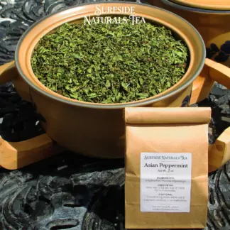 Tin-tie kraft package sitting on a black wrought-iron table with a white label that shows the brand (Surfside Naturals Tea), product name (Asian Peppermint, Net Wt. 2 oz), ingredients (Wildcrafted Mentha haplocalyx), directions (Slow boil 1 tsp per cup of water for 3-5 minutes), and cautions (Consult a physician before use if pregnant, nursing, or on medication). A large tan Pfaltzgraff Folk Art stoneware bowl filled with a mix of clean, bright, and dark green mint leaves sits behind the package.