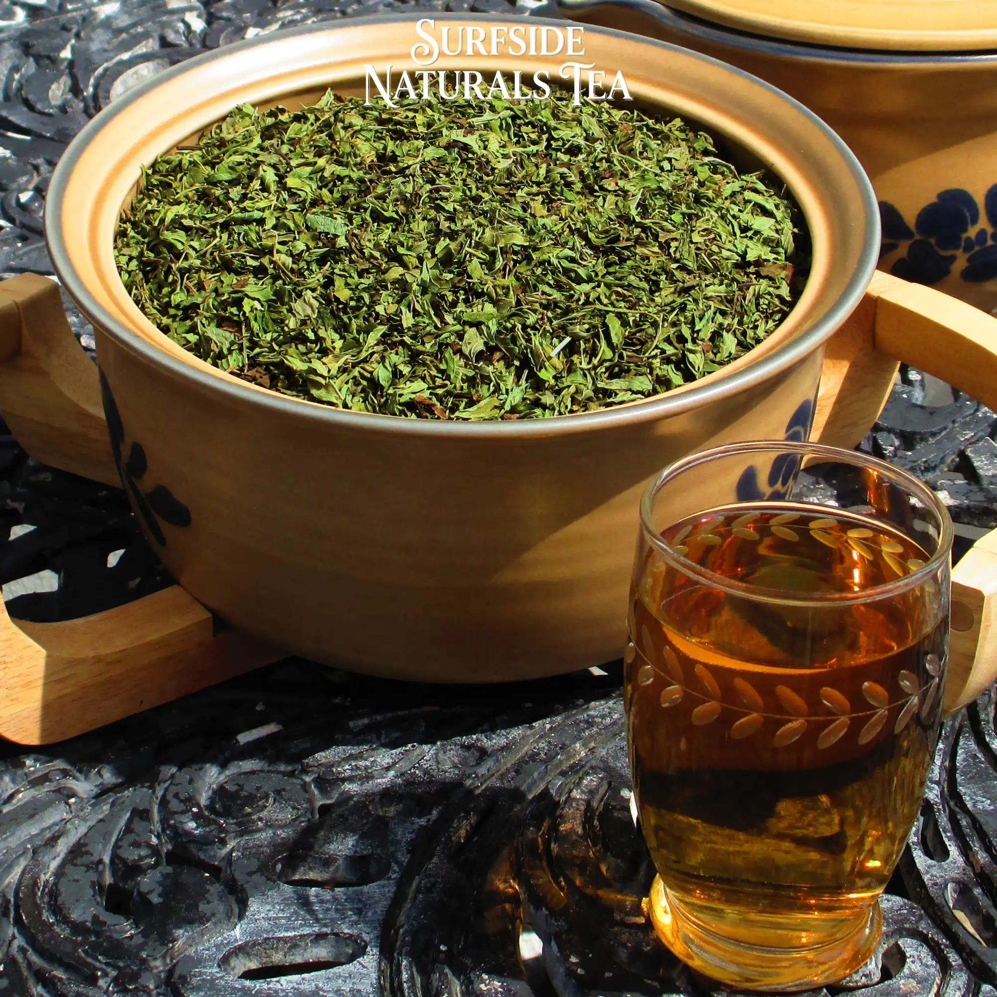 Tan Pfaltzgraff Folk Art stoneware bowl sitting in a wooden serving cradle, filled with dried Asian Peppermint (a mix of clean, bright, and dark green mint leaves), on a black wrought-iron table, with a glass of amber tea.