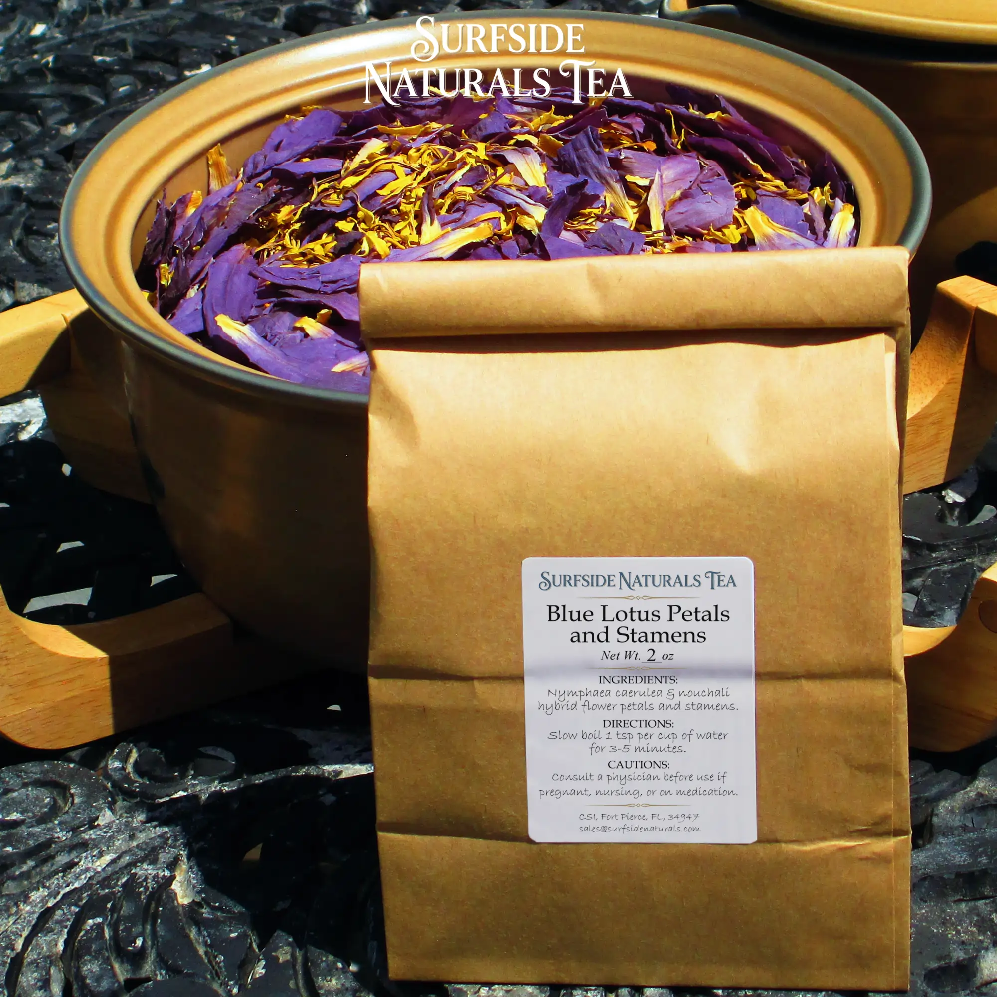 Tin-tie kraft package sitting on a black wrought-iron table with a white label that shows the brand (Surfside Naturals Tea), product name (Blue Lotus Petals and Stamens, Net Wt. 2 oz), ingredients (Nymphaea caerulea & nouchali hybrid flower petals and stamens), directions (Slow boil 1 tsp per cup of water for 3-5 minutes), and cautions (Consult a physician before use if pregnant, nursing, or on medication). A large tan Pfaltzgraff Folk Art stoneware bowl filled with dried blue lotus petals and stamens (clean, fresh light-to-dark purple petals with bright yellow stamens) sits behind the package.
