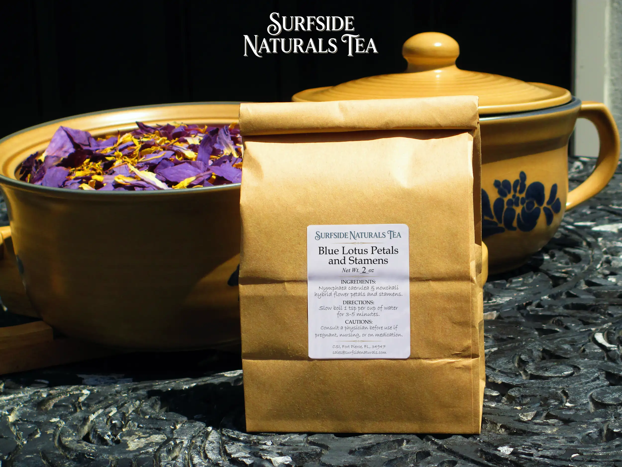 Tin-tie kraft package sitting on a black wrought-iron table with a white label that shows the brand (Surfside Naturals Tea), product name (Blue Lotus Petals and Stamens, Net Wt. 2 oz), ingredients (Nymphaea caerulea & nouchali hybrid flower petals and stamens), directions (Slow boil 1 tsp per cup of water for 3-5 minutes), and cautions (Consult a physician before use if pregnant, nursing, or on medication). Behind the package, a tan Pfaltzgraff Folk Art stoneware bowl filled with dried blue lotus petals and stamens (clean, fresh light-to-dark purple petals with bright yellow stamens) is to the left, and a matching pot with a lid is to the right.