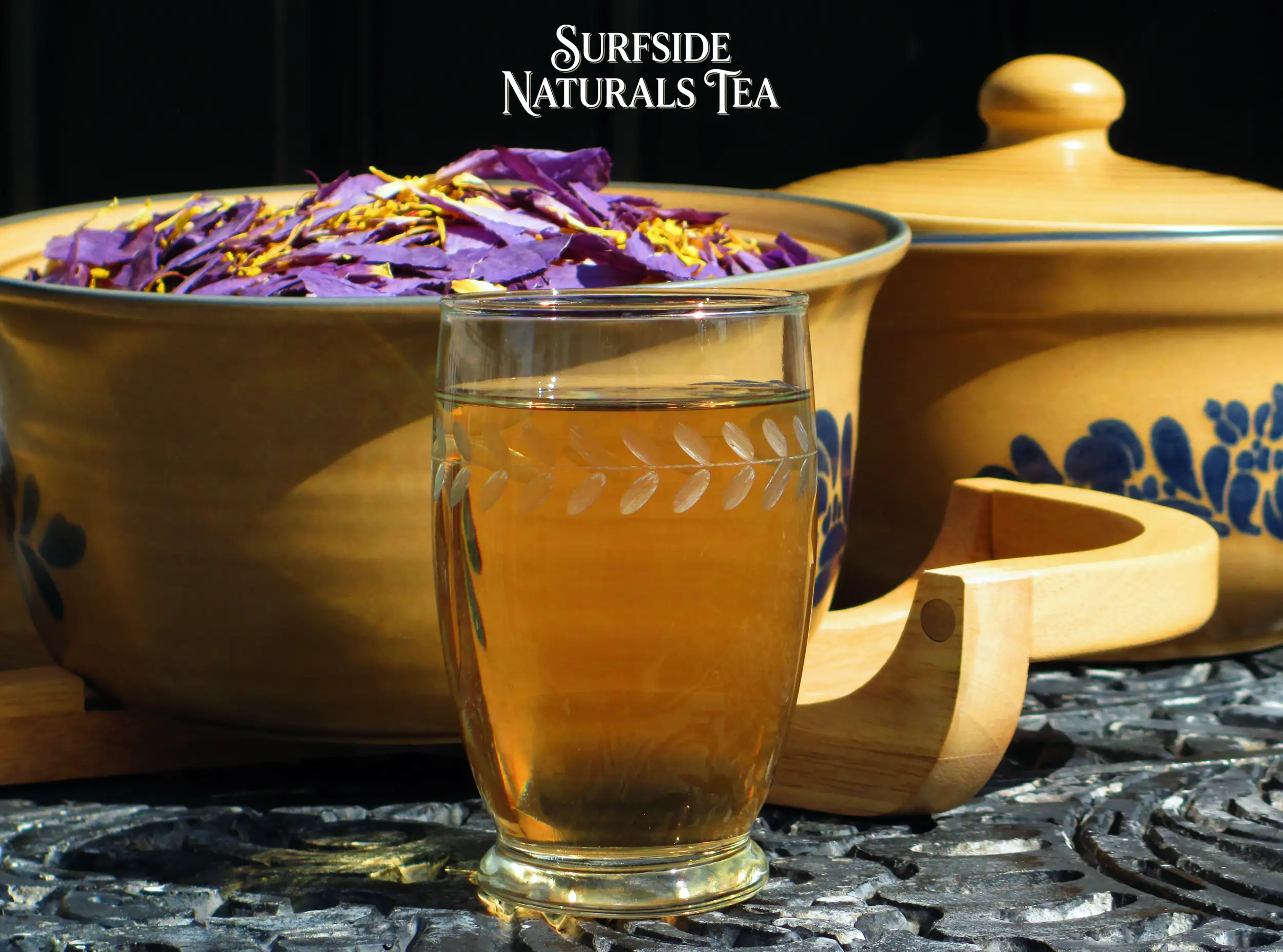 Blue lotus tea in a clear leaf-patterned glass, sitting on a black wrought-iron table. Behind it is a Pfaltzgraff Folk Art stoneware bowl filled with dried blue lotus petals. A small Surfside Naturals Tea logo is centered at the top.