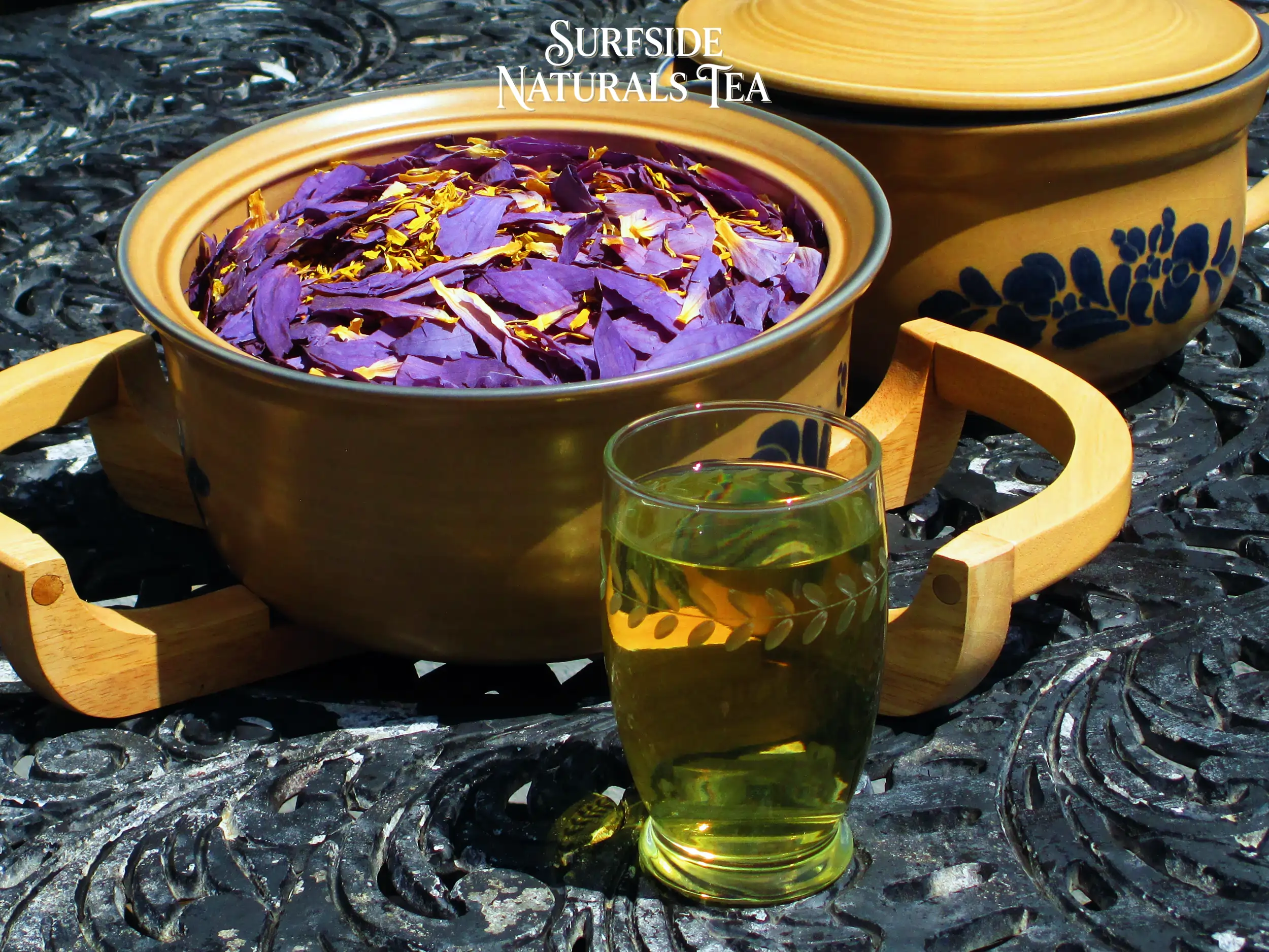 Tan Pfaltzgraff Folk Art stoneware bowl sitting in a wooden serving cradle, filled with dried Blue Lotus petals and stamens (clean, light-to-dark purple petals with bright yellow stamens), on a black wrought-iron table. A matching lidded pot is partially visible to the right, with a glass of blueish yellow tea in front. A small Surfside Naturals Tea logo is at the top of the image.