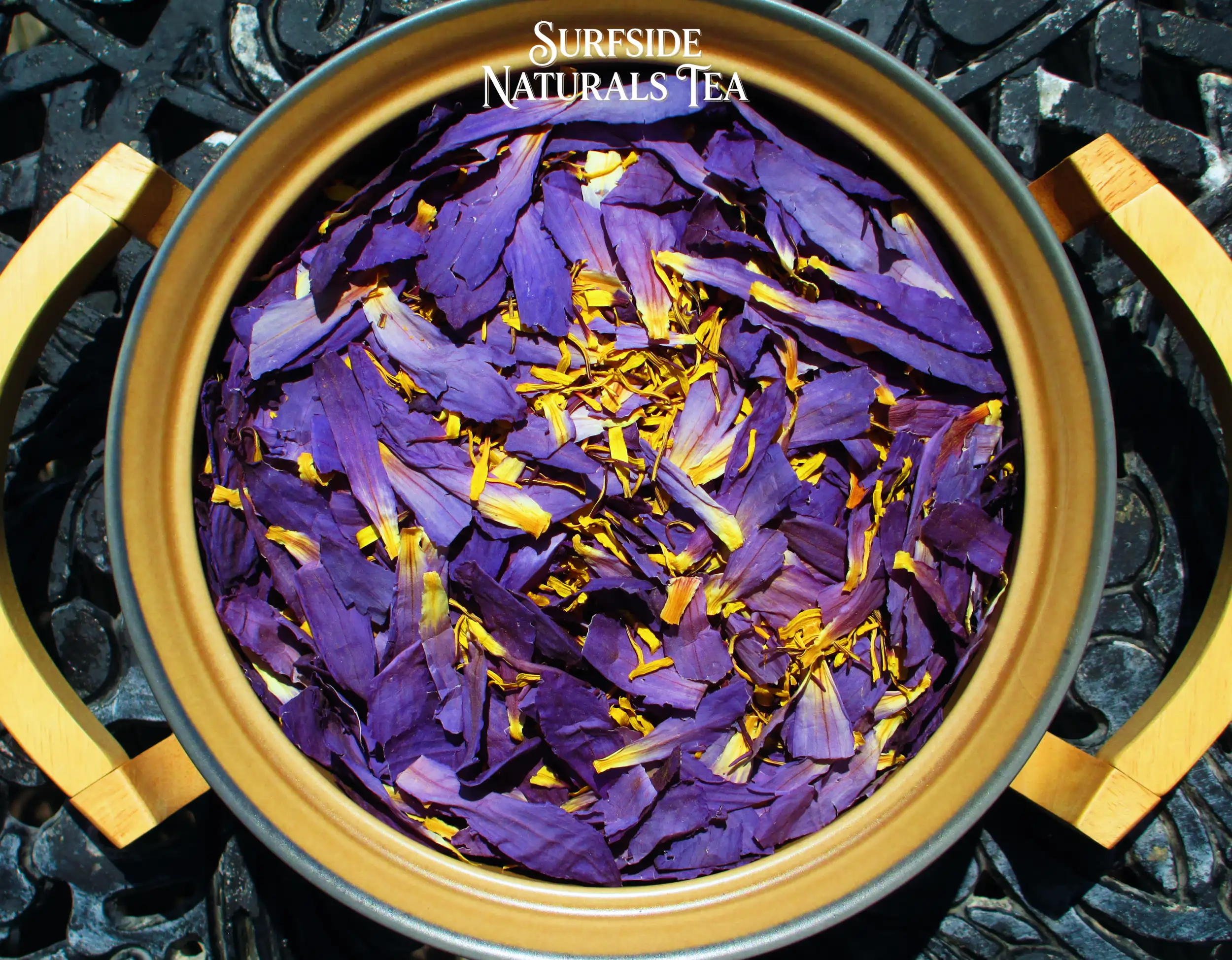 Top-down view of a tan Pfaltzgraff Folk Art stoneware bowl sitting in a wooden serving cradle, filled with dried blue lotus petals and stamens (clean, fresh light-to-dark purple petals with bright yellow stamens), resting on a black wrought-iron table.