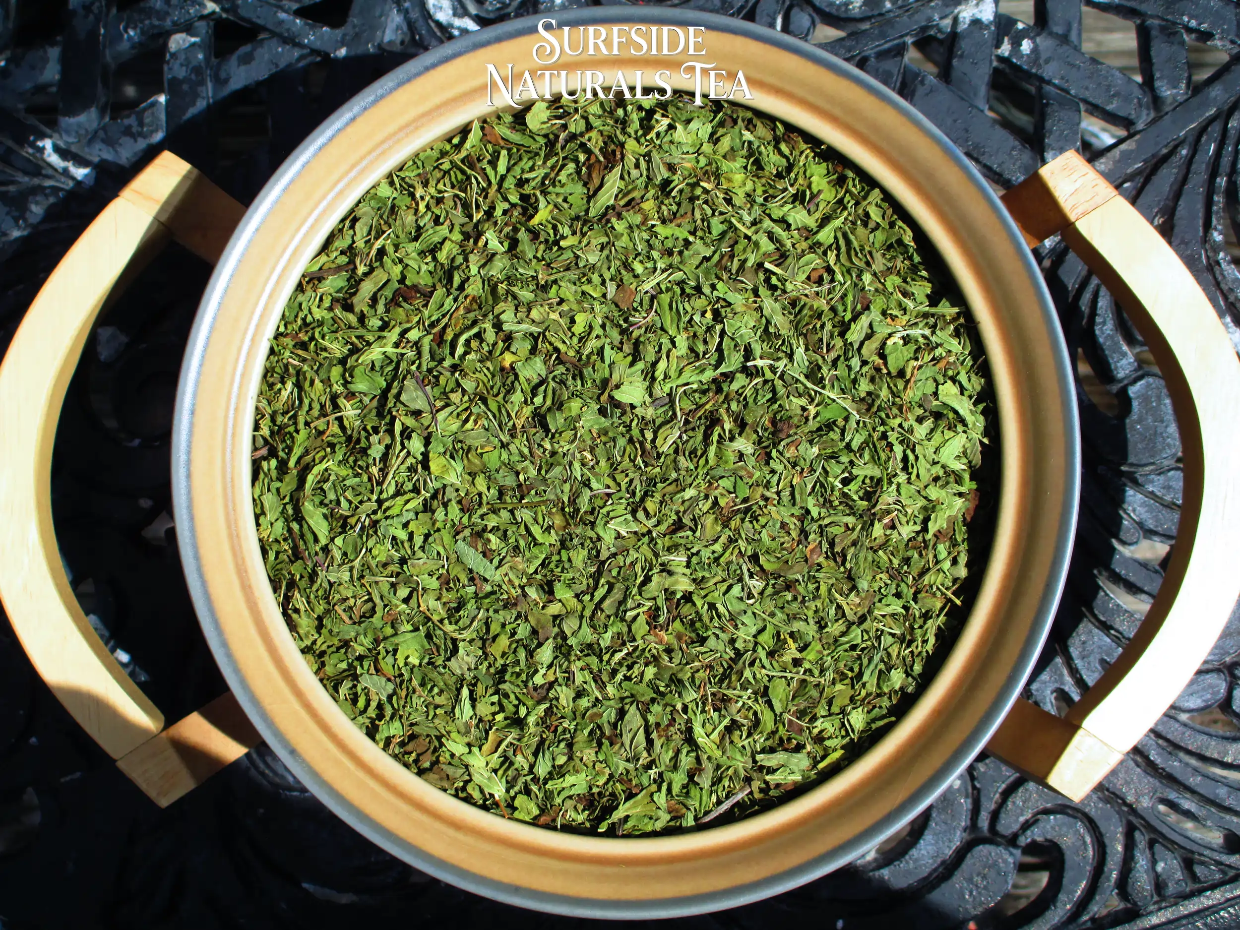 Top-down view of a tan Pfaltzgraff Folk Art stoneware bowl sitting in a wooden serving cradle, filled with dried Asian Peppermint (a mix of clean bright and dark green mint leaves), resting on a black wrought-iron table.