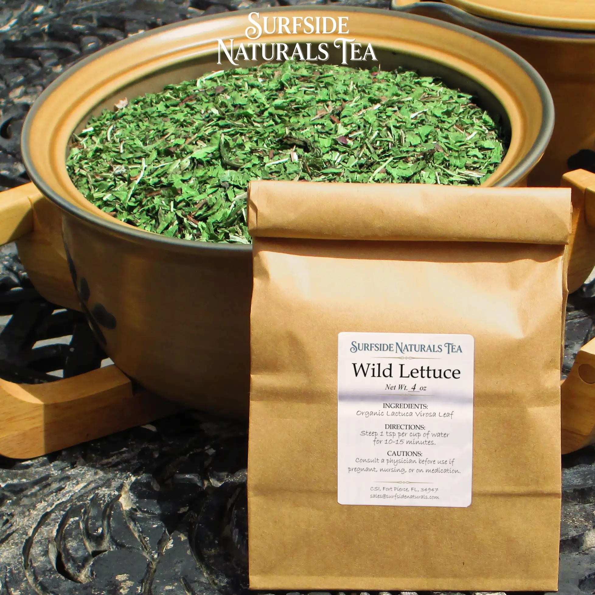Tin-tie kraft package sitting on a black wrought-iron table with a white label that shows the brand (Surfside Naturals Tea), product name (Wild Lettuce, Net Wt. 4 oz), ingredients (Organic Lactuca Virosa Leaf), directions (Steep 1 tsp per cup of water for 10–15 minutes), and cautions (Consult a physician before use if pregnant, nursing, or on medication). A large tan Pfaltzgraff Folk Art stoneware bowl filled with clean, light green pieces of wild lettuce leaf sits behind the package.