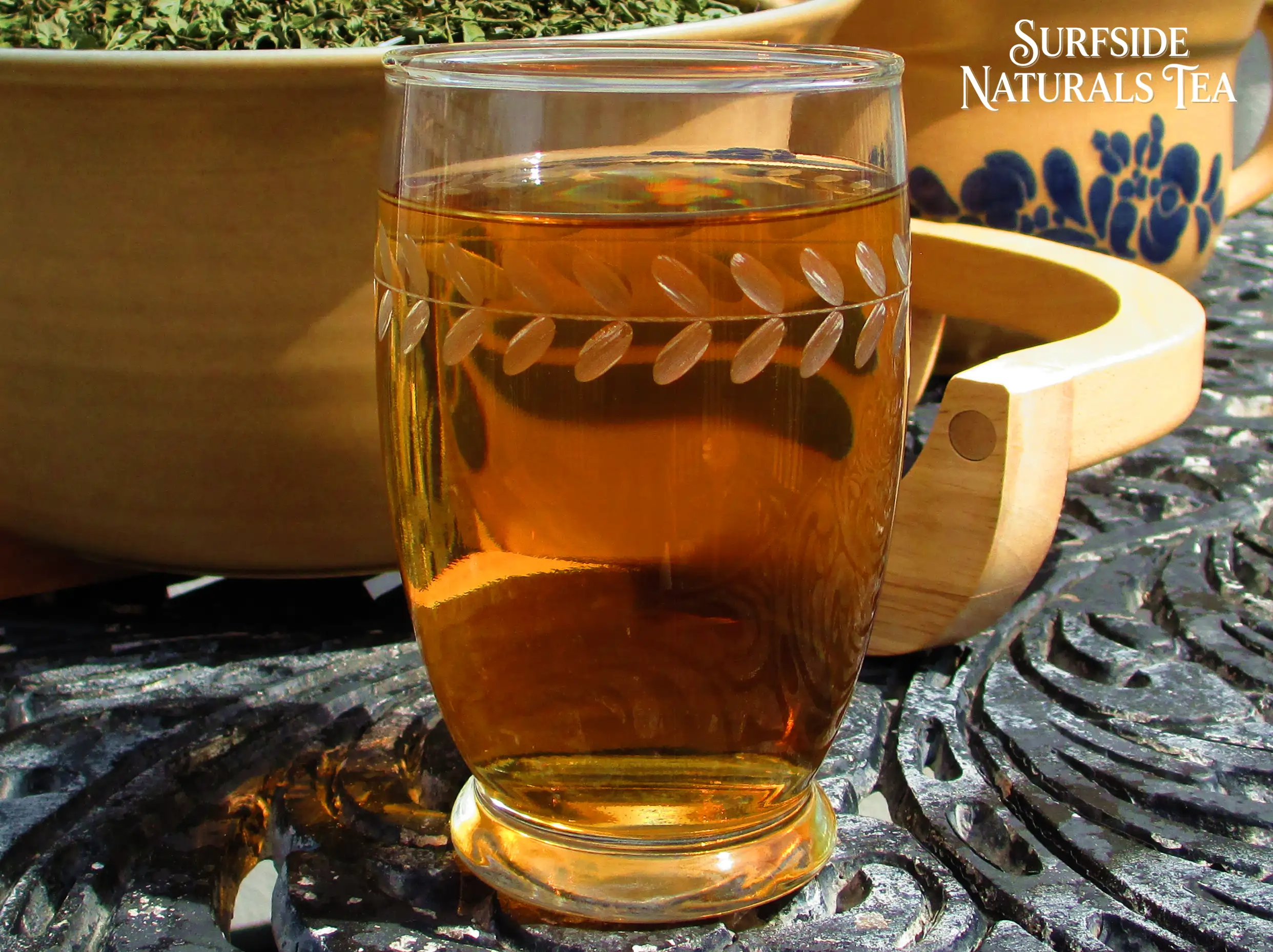 Asian Peppermint tea (amber in color) in a clear leaf-patterned glass, sitting on a black wrought-iron table. Behind it is a Pfaltzgraff Folk Art stoneware bowl filled with dried Asian Peppermint leaves.