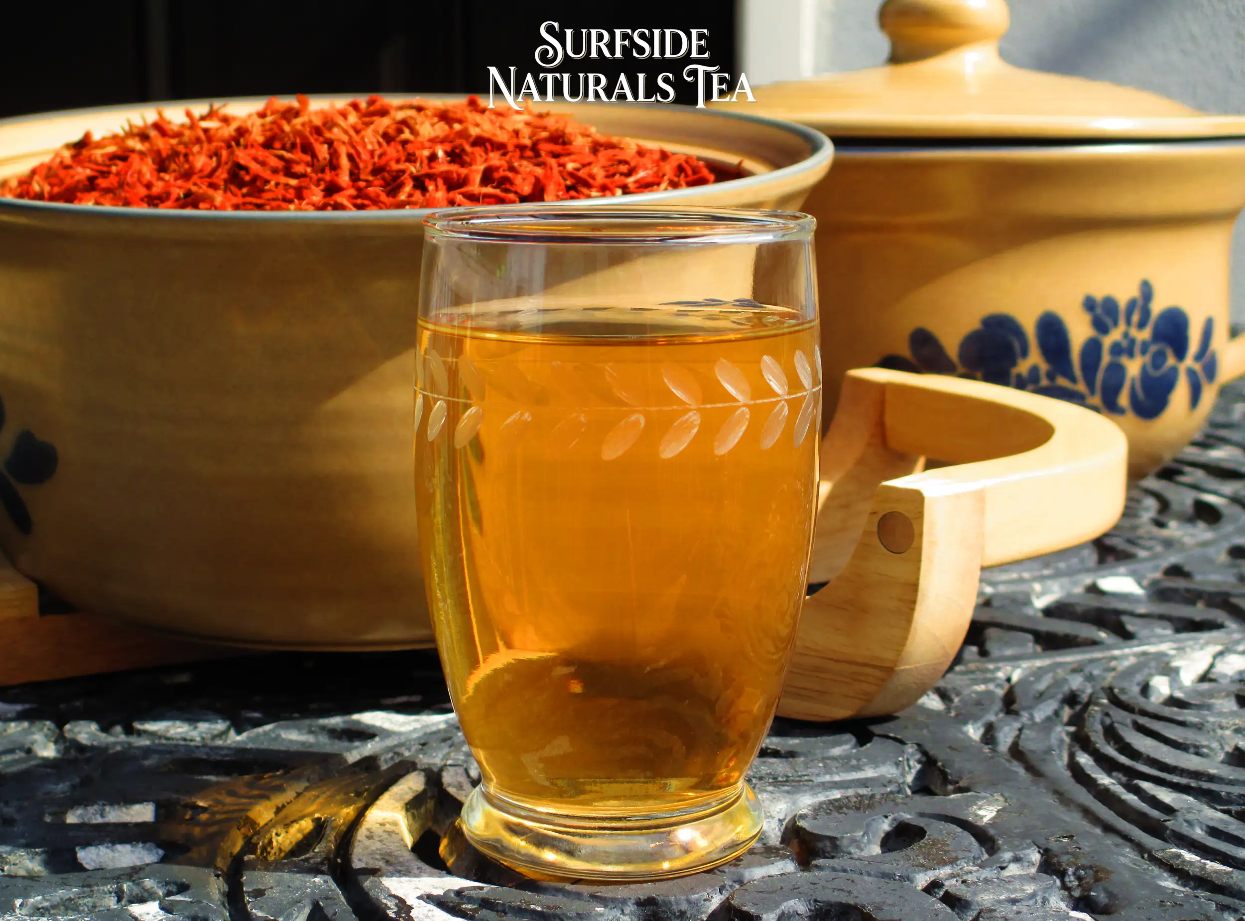 Klip dagga tea in a clear leaf-patterned glass, sitting on a black wrought-iron table. Behind it is a Pfaltzgraff Folk Art stoneware bowl filled with bright red klip dagga petals.