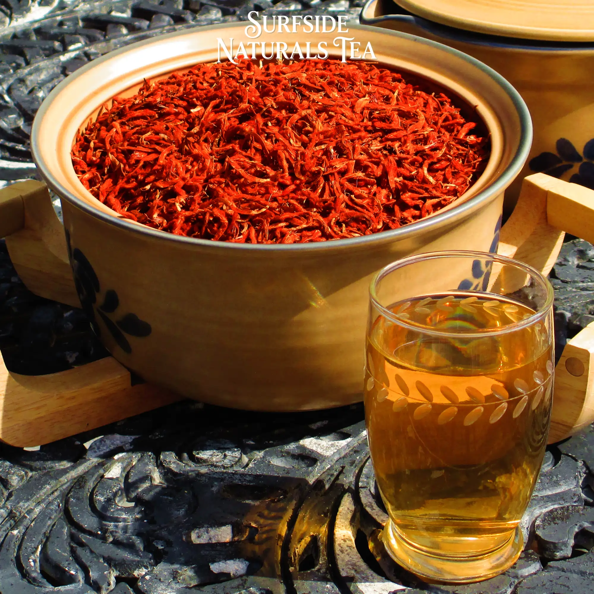 Tan Pfaltzgraff Folk Art stoneware bowl sitting in a wooden serving cradle, filled with dried klip dagga flowers (clean bright red, whole petals), on a black wrought-iron table. A matching lidded pot is partially visible to the right, with a glass of golden tea in front.