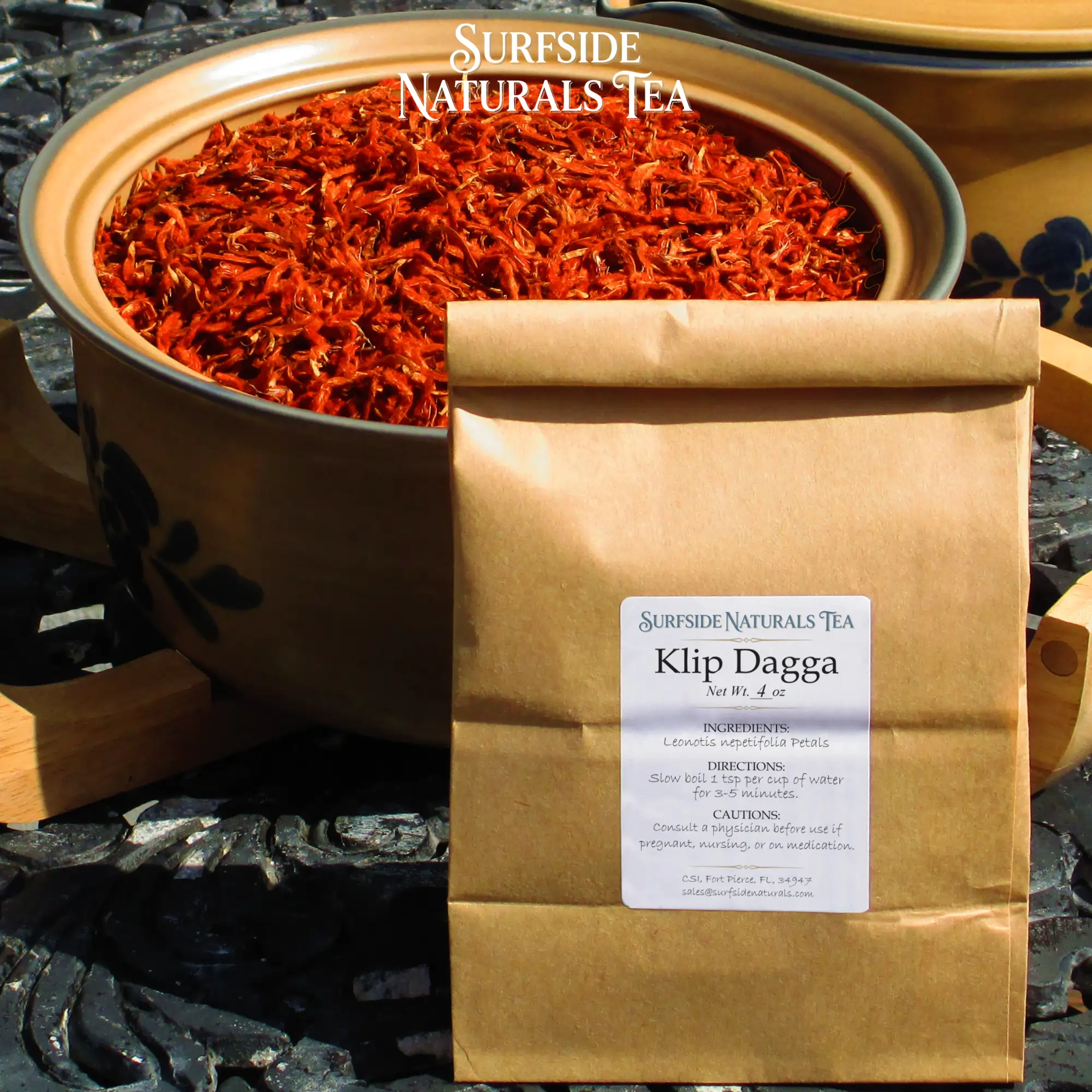 Tin-tie kraft package sitting on a black wrought-iron table with a white label that shows the brand (Surfside Naturals Tea), product name (Klip Dagga, Net Wt. 4 oz), ingredients (Leonotis nepetifolia Petals), directions (Slow boil 1 tsp per cup of water for 3-5 minutes), and cautions (Consult a physician before use if pregnant, nursing, or on medication). A large tan Pfaltzgraff Folk Art stoneware bowl filled with clean, bright red klip dagga petals sits behind the package.