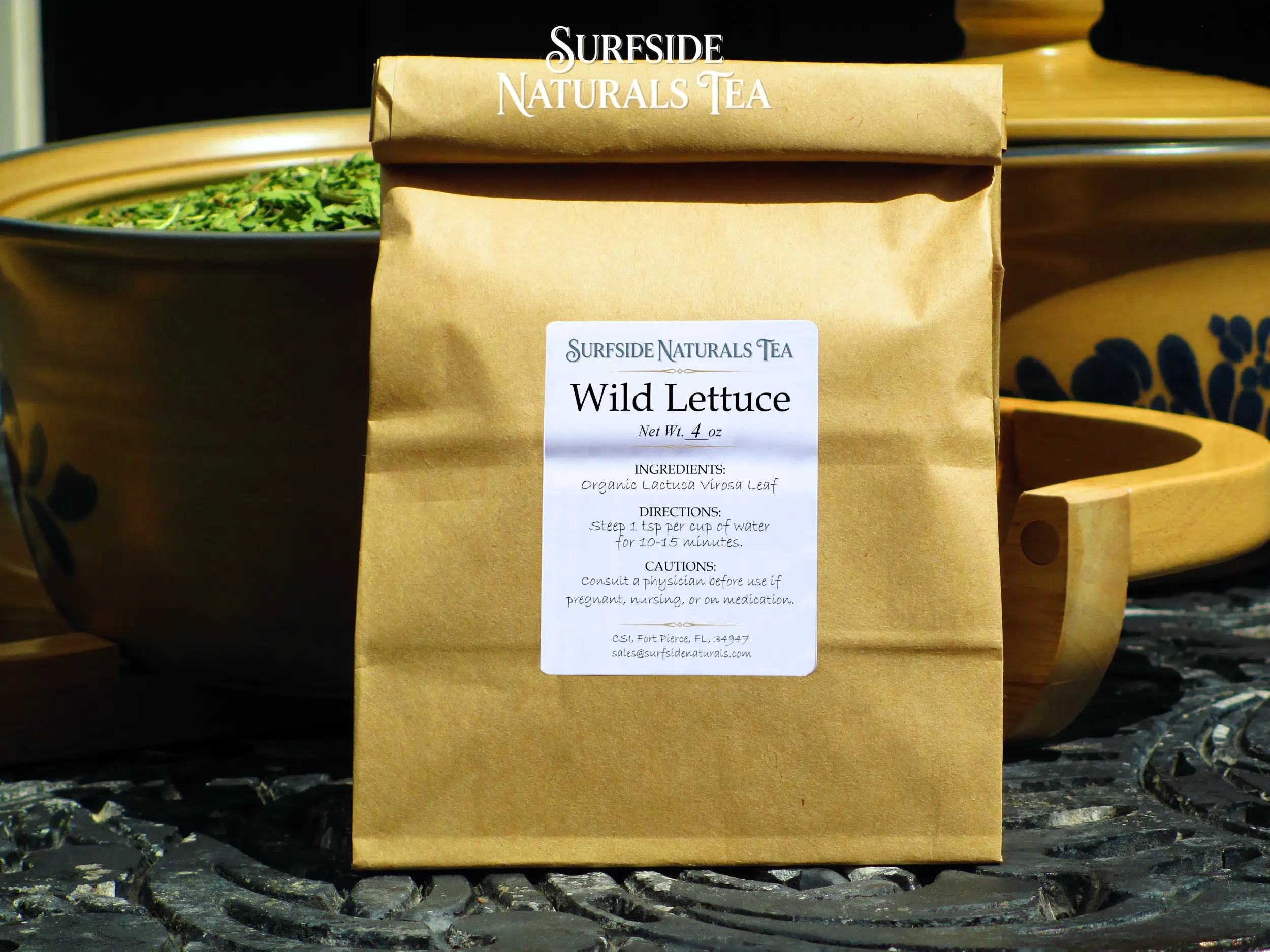 Tin-tie kraft package sitting on a black wrought-iron table with a white label that shows the brand (Surfside Naturals Tea), product name (Wild Lettuce, Net Wt. 4 oz), ingredients (Organic Lactuca Virosa Leaf), directions (Slow boil 1 tsp per cup of water for 3-5 minutes), and cautions (Consult a physician before use if pregnant, nursing, or on medication). Behind the package, a tan Pfaltzgraff Folk Art stoneware bowl filled with light green pieces of wild lettuce leaf and a matching pot with a lid is to the right.